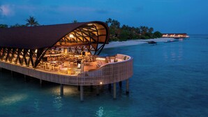 An overwater resort with a covered dining area, lounge chairs, and a view of the beach and palm trees. - The Westin Maldives Miriandhoo Resort (Miriandhoo Island)