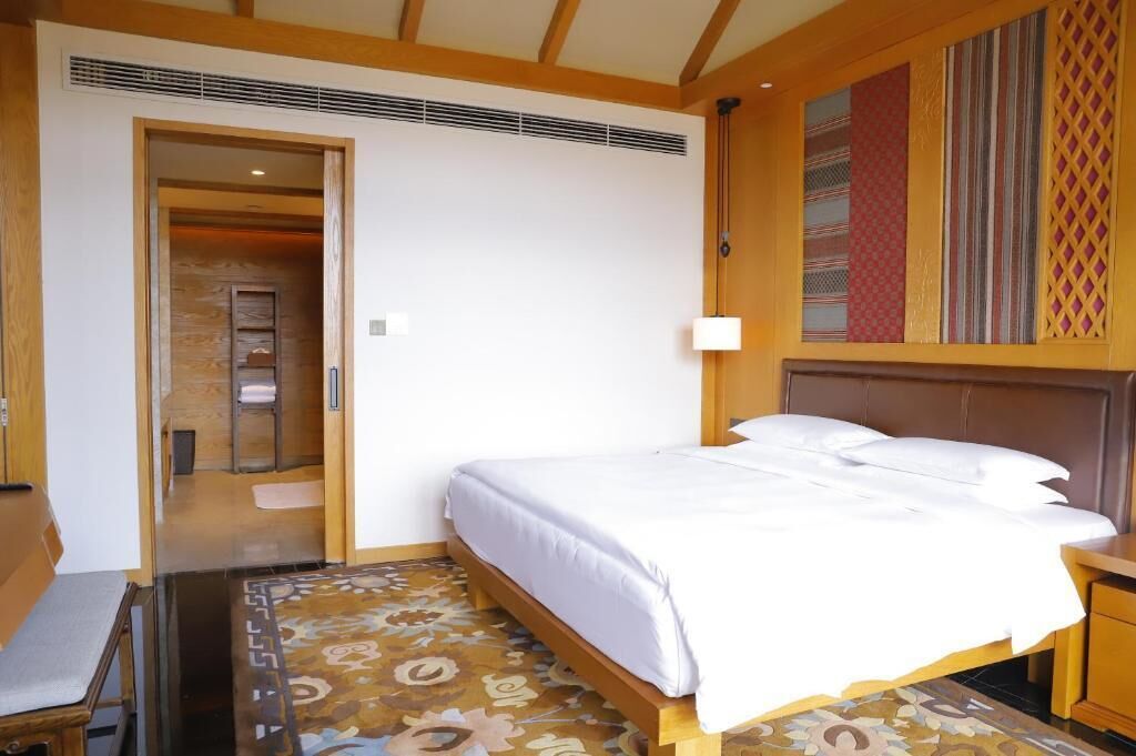 Ein Hotelzimmer mit Bett, Nachttisch, Lampe, einem gemusterten Teppich und einer Tür zu einem weiteren Raum.