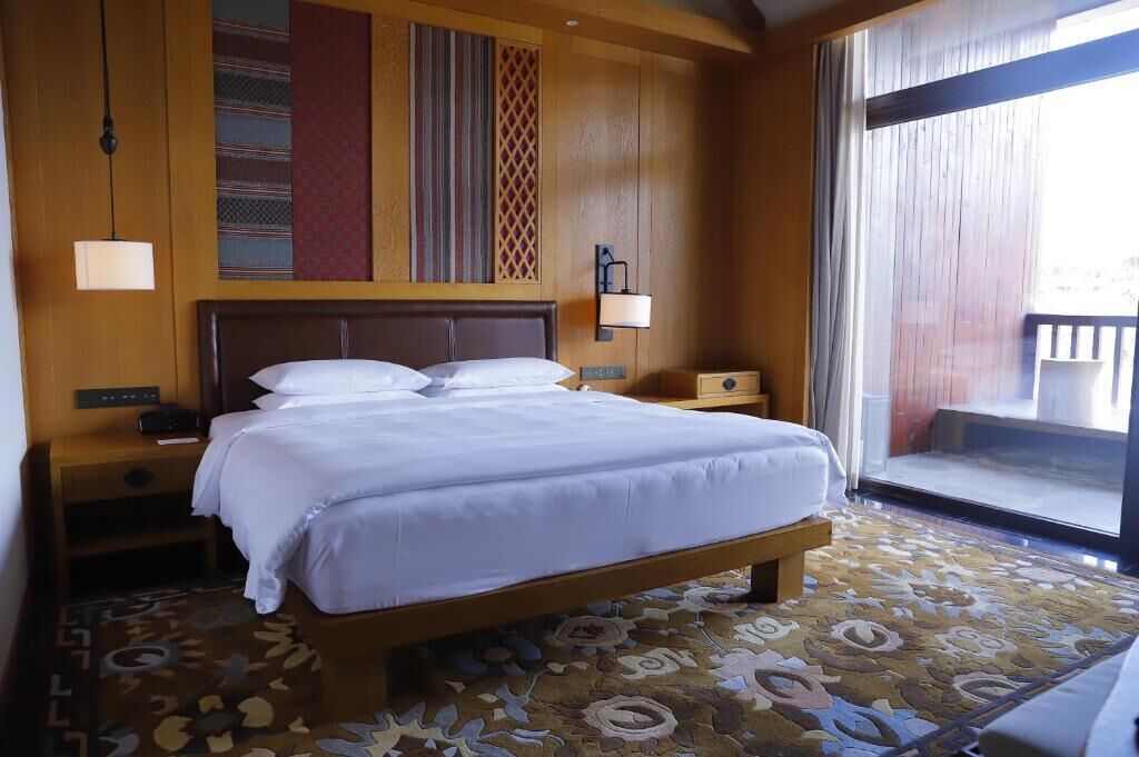 A hotel room with a large bed, a nightstand, a lamp, and a view of the outside.
