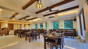 A spacious dining area with high ceilings, wooden beams, and large windows offering views of greenery. Tables are set with chairs, and pendant lights hang above. - Hukam's Holiday Home (Bhuntar)