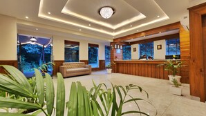 A hotel lobby with a reception desk, a sofa, and large windows offering a view of the outdoors. - Hukam's Holiday Home (Bhuntar)