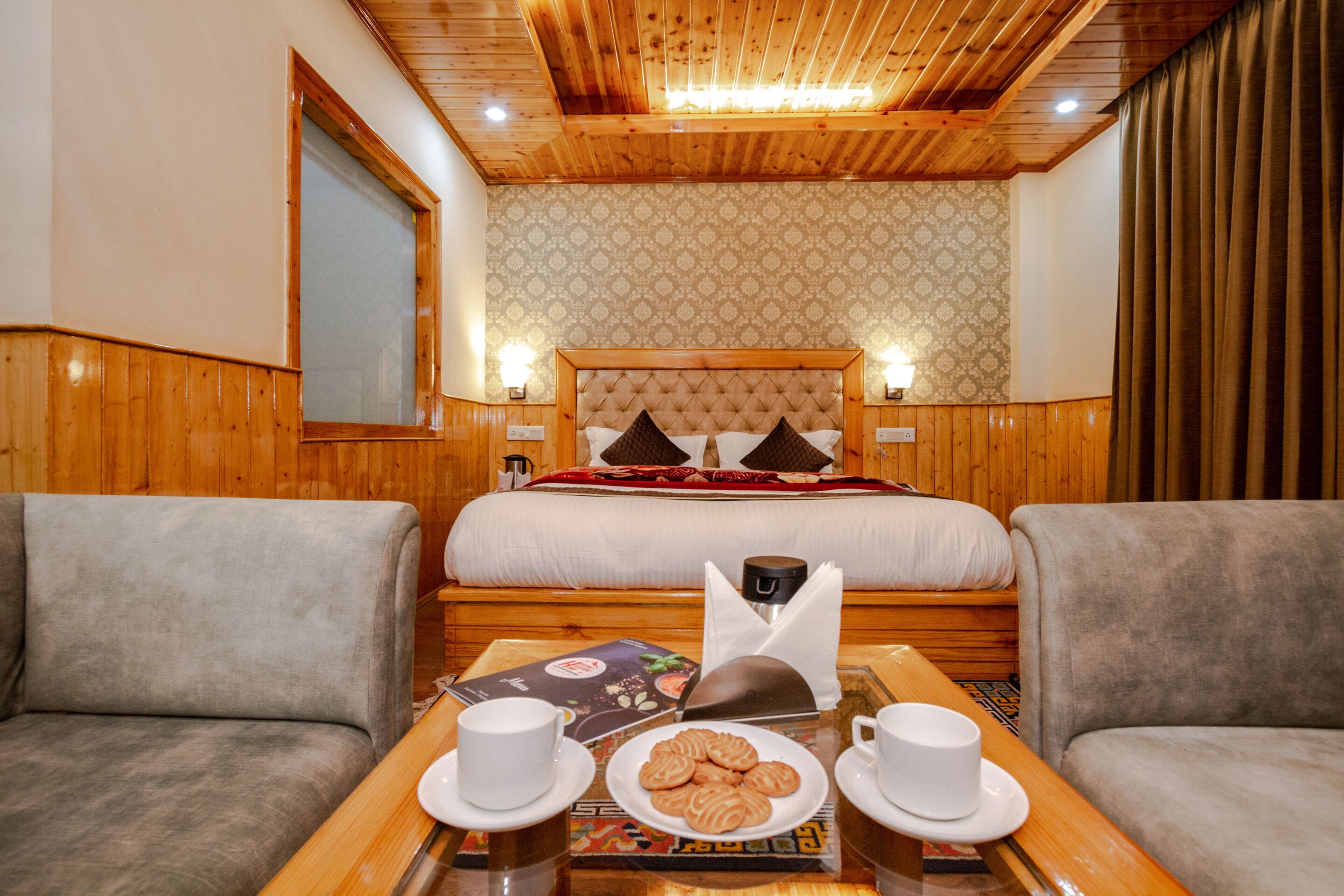 A hotel room with a wooden ceiling, a bed with a tufted headboard, two armchairs, a table with a plate of cookies, two cups, and a menu.