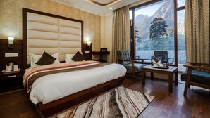A hotel room with a large bed, a view of mountains, a sitting area with a table and chairs, and a window with curtains. - Hukam's Holiday Home (Bhuntar)
