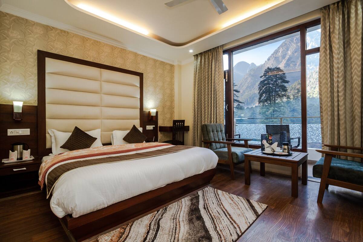 Quarto de hotel com uma cama grande, vista para montanhas, área de estar com mesa e cadeiras, e janela com cortinas.