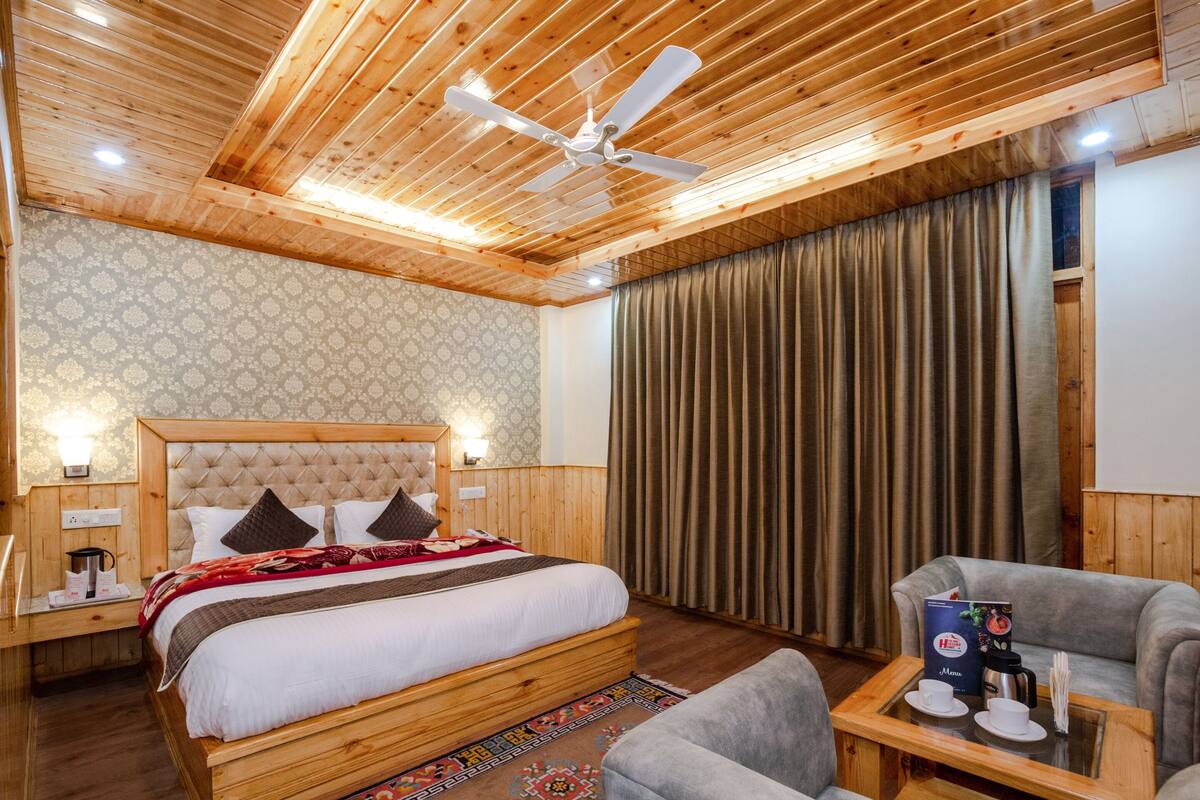 Quarto de hotel com teto de madeira, cama com cabeceira estofada, poltrona cinza, mesa de cabeceira de madeira com um menu e uma janela grande com cortinas.
