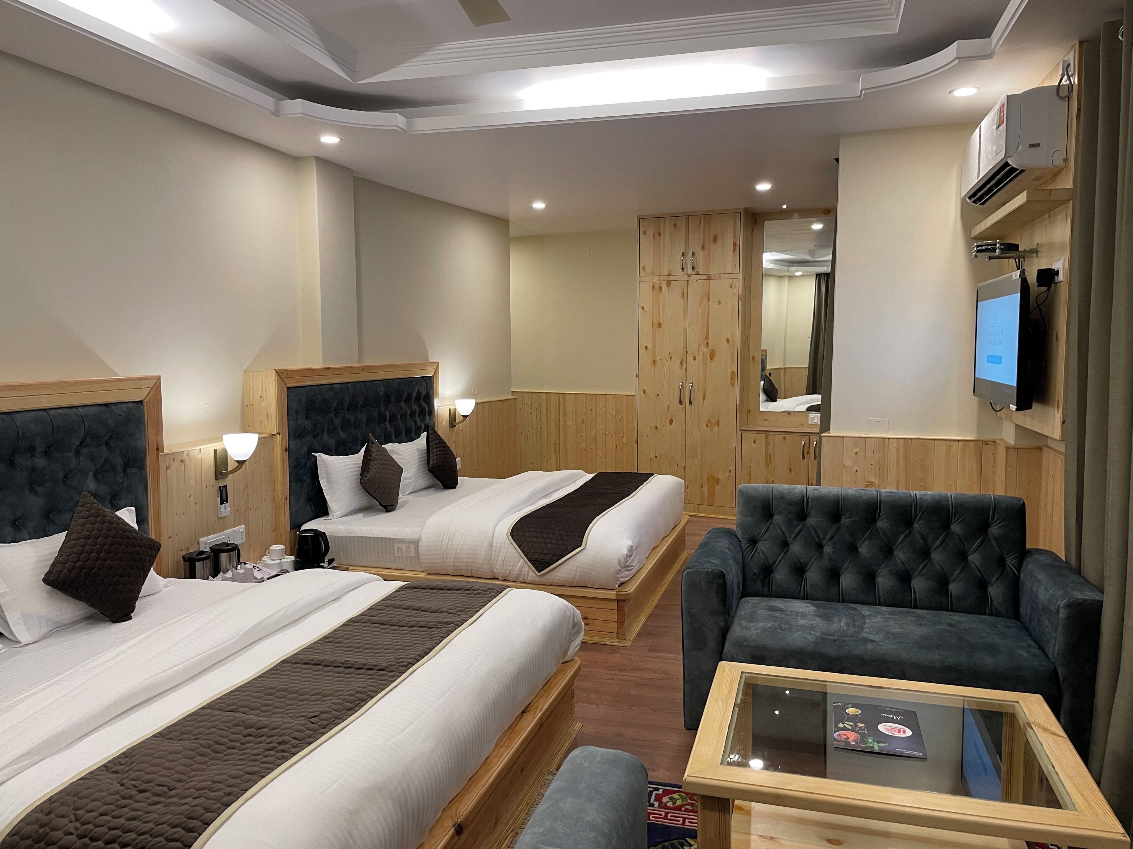 Habitación de hotel con dos camas, un sillón gris, una mesita, armario de madera y televisor montado en la pared.
