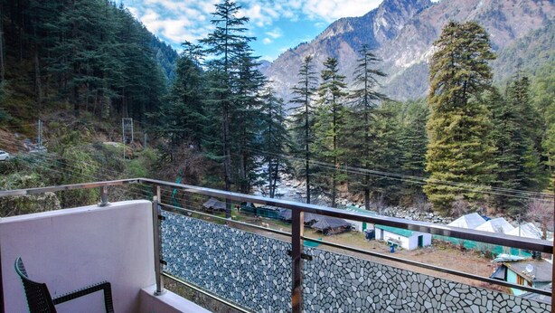 A balcony with a wooden table and chairs, offering a view of a mountainous landscape with trees and a road. - Hukam's Holiday Home (Bhuntar)