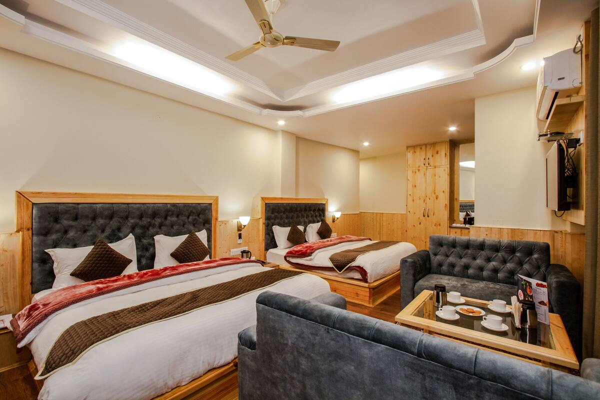 Quarto de hotel com duas camas, um sofá, uma mesa de vidro com chávenas e um menu, e um ventilador de teto.