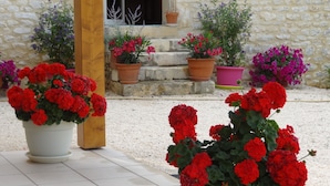 Une cour avec des géraniums et d’autres fleurs en pot, devant un bâtiment en pierre.