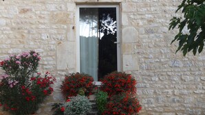 Un bâtiment en pierre doté d’une fenêtre et d’une porte, entouré de plantes en pot ornées de fleurs rouges et roses.