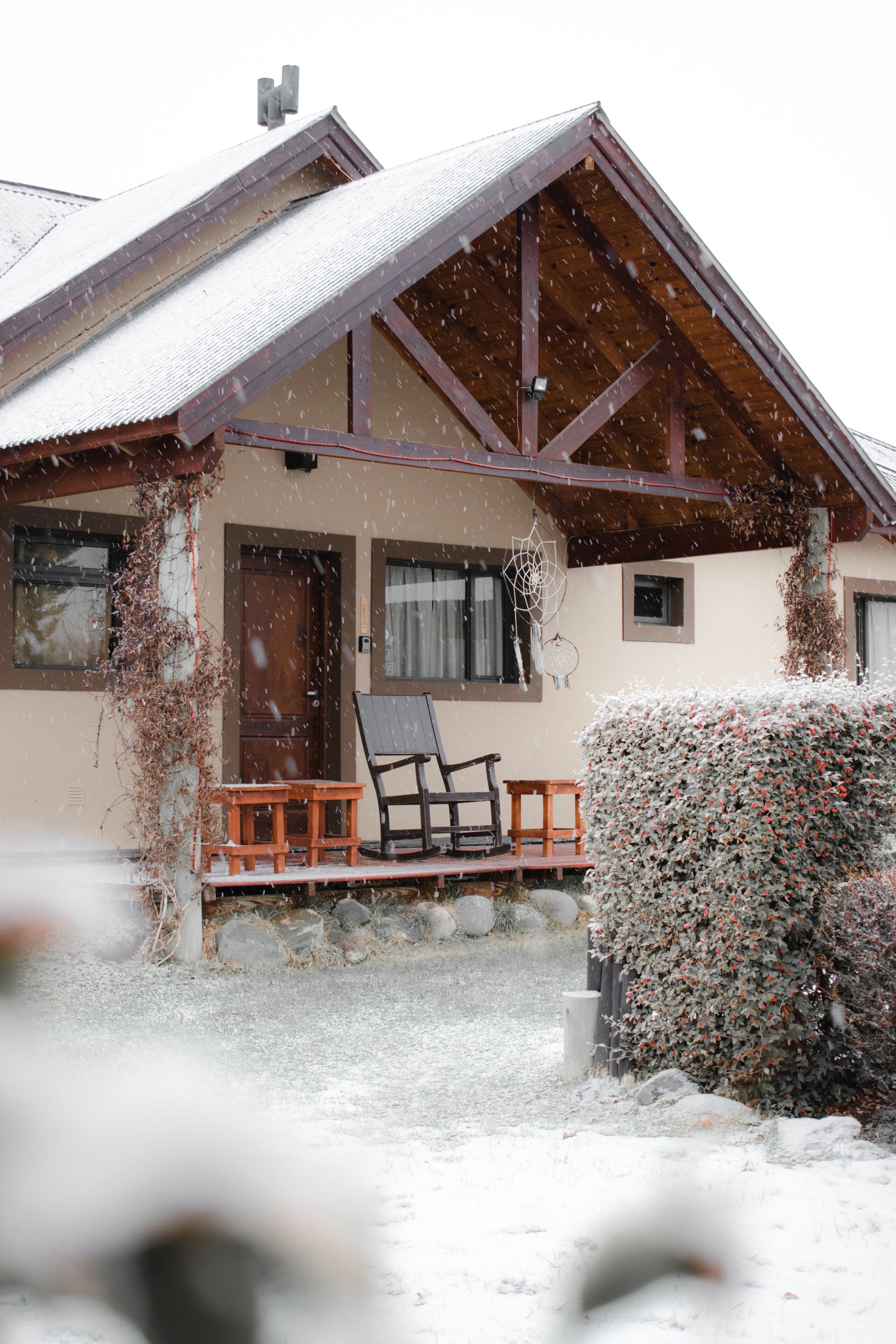 A wooden house with a sloped roof, a porch with two chairs, and a snow-covered ground.