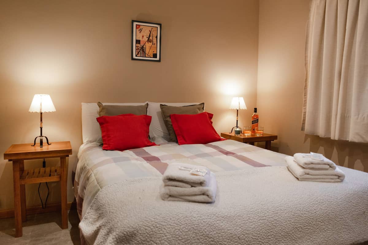 Een hotelkamer met een bed, twee nachtkastjes, lampen, een lijst met een afbeelding aan de muur en een raam met gordijnen.