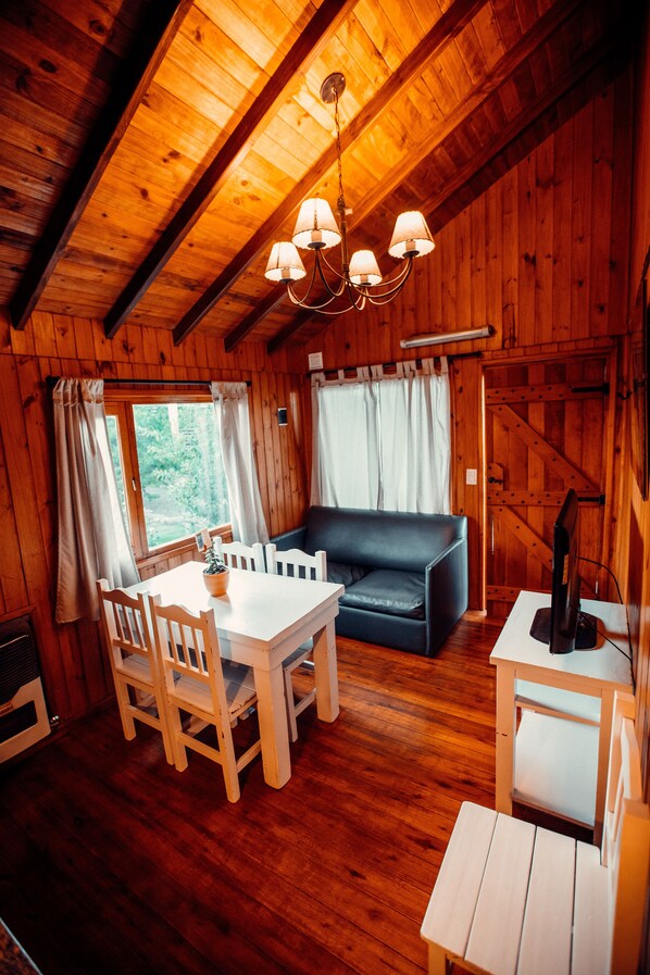 A wooden cabin interior with a dining table, chairs, a sofa, and a television.