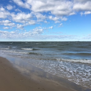 Une plage avec des vagues, une jetée visible au loin et un ciel partiellement nuageux.