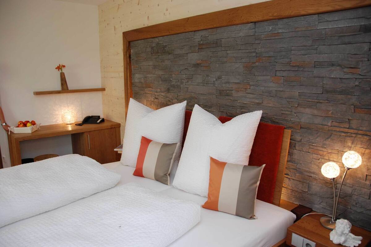 Uma cama bem arrumada com roupa de cama branca e almofadas listradas, uma mesinha de cabeceira de madeira com abajur, e uma parede de pedra ao fundo.