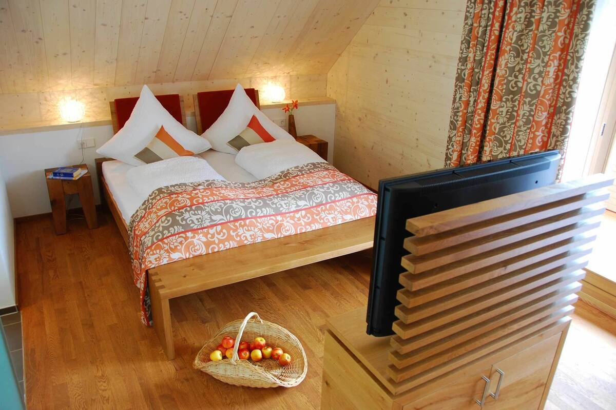 Uma cama de madeira com colcha estampada, uma mesinha de cabeceira com um livro e uma cesta de maçãs no chão.