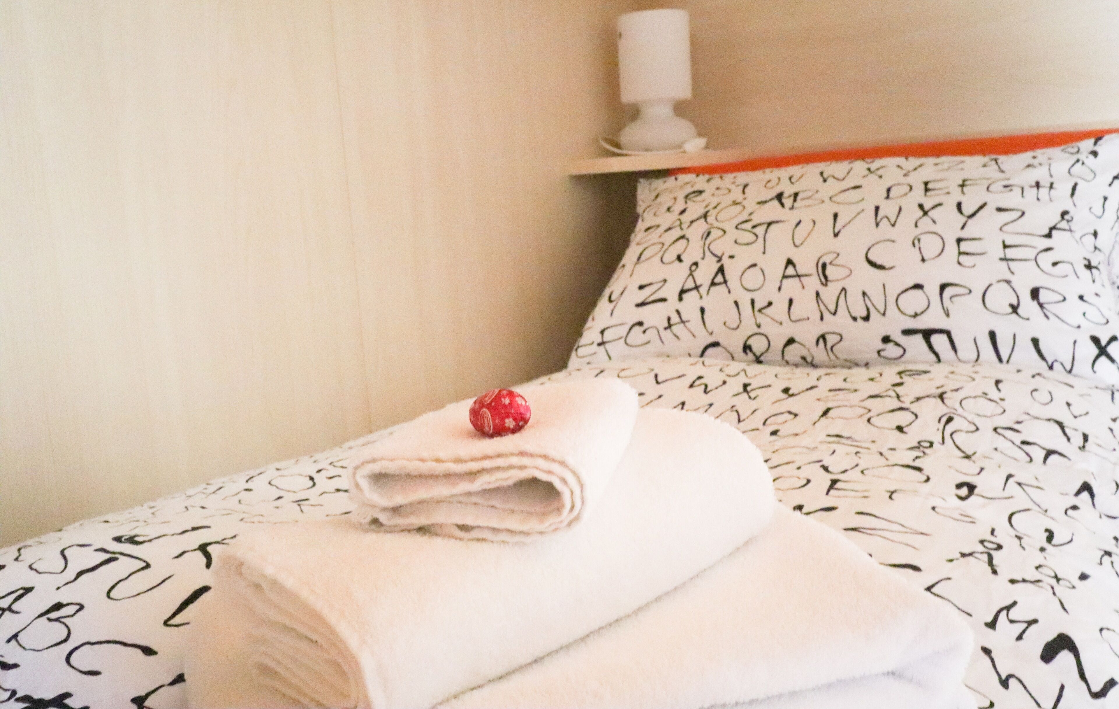 Een net opgemaakt bed met een zwart-wit kussenhoes met een alfabetpatroon, een opgevouwen witte handdoek en een rode decoratie op het bed.