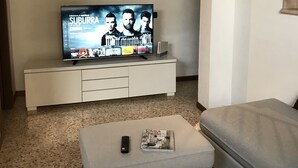 Un salon avec un téléviseur à écran plat posé sur un meuble de télévision blanc, une télécommande et un magazine sur un pouf gris.