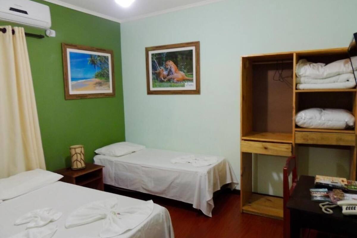 Habitación de hotel con dos camas individuales, un armario de madera, una mesita con artículos de aseo y dos cuadros en la pared.