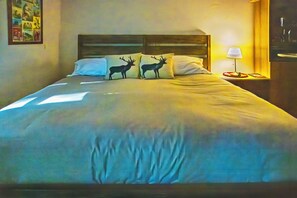 A bed with a headboard, two pillows with deer silhouettes, a bedside table with a lamp, and a wall with a framed picture.