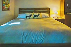 Una cama con cabecera, dos almohadas con siluetas de ciervo, una mesita de noche con una lámpara y una pared con un cuadro enmarcado.