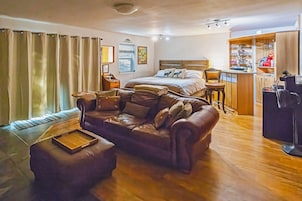 Una habitación acogedora con un sofá de cuero, una cama con almohadones decorativos, piso de madera, una mesita y una pequeña zona de kitchenette.