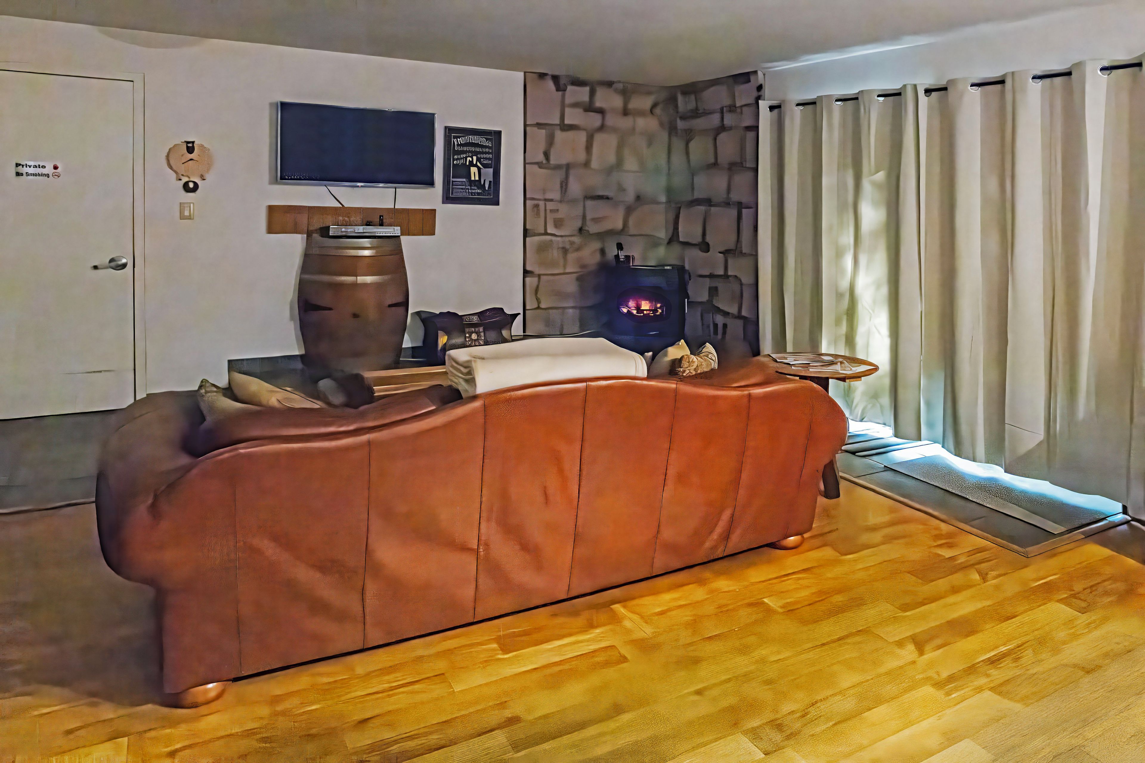Un salon avec un canapé marron, un téléviseur fixé au mur, un mur en pierre et une fenêtre avec des rideaux.