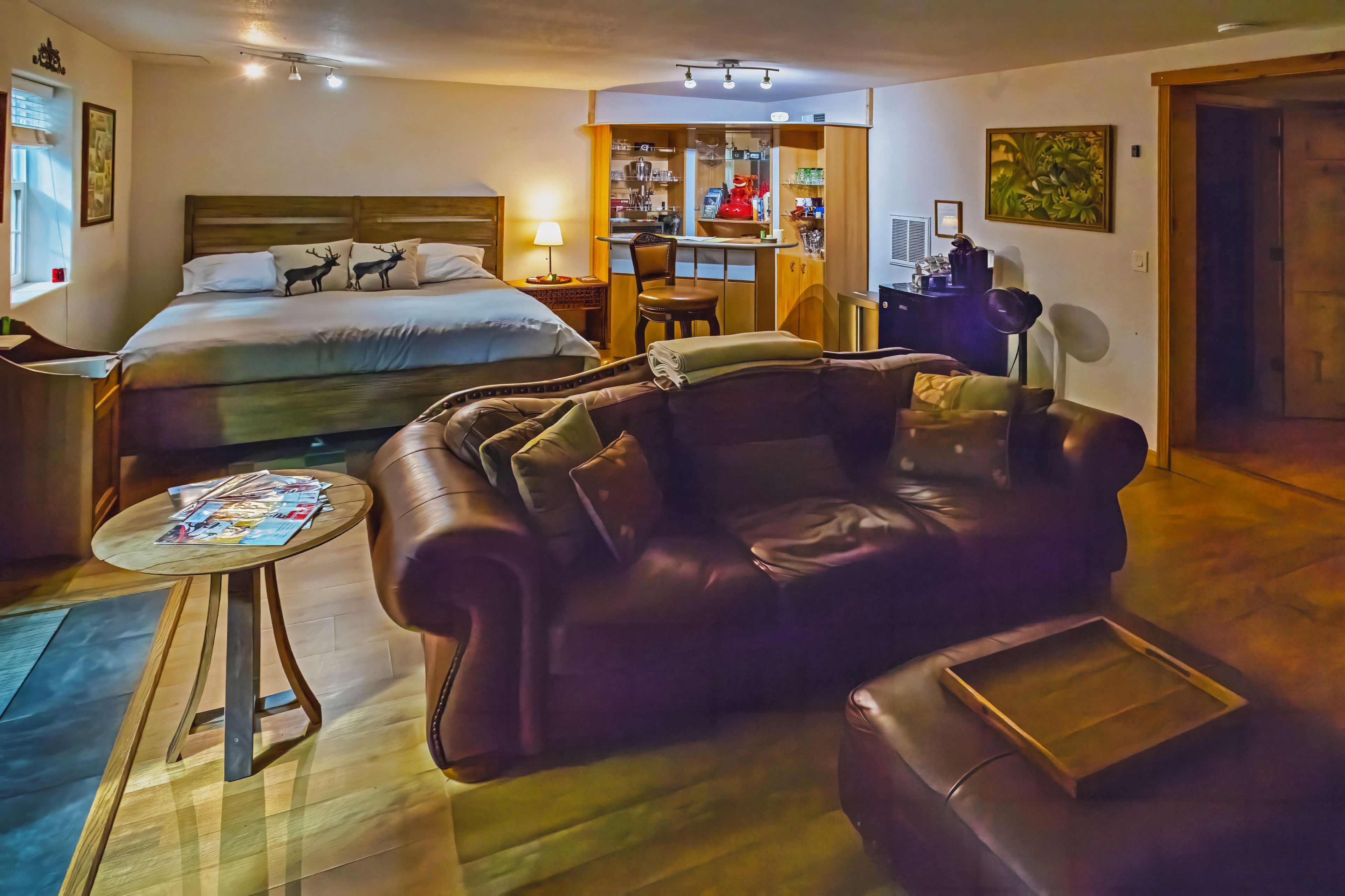 A hotel room with a bed, a leather sofa, a small table with magazines, a kitchenette, and a door leading to another room.