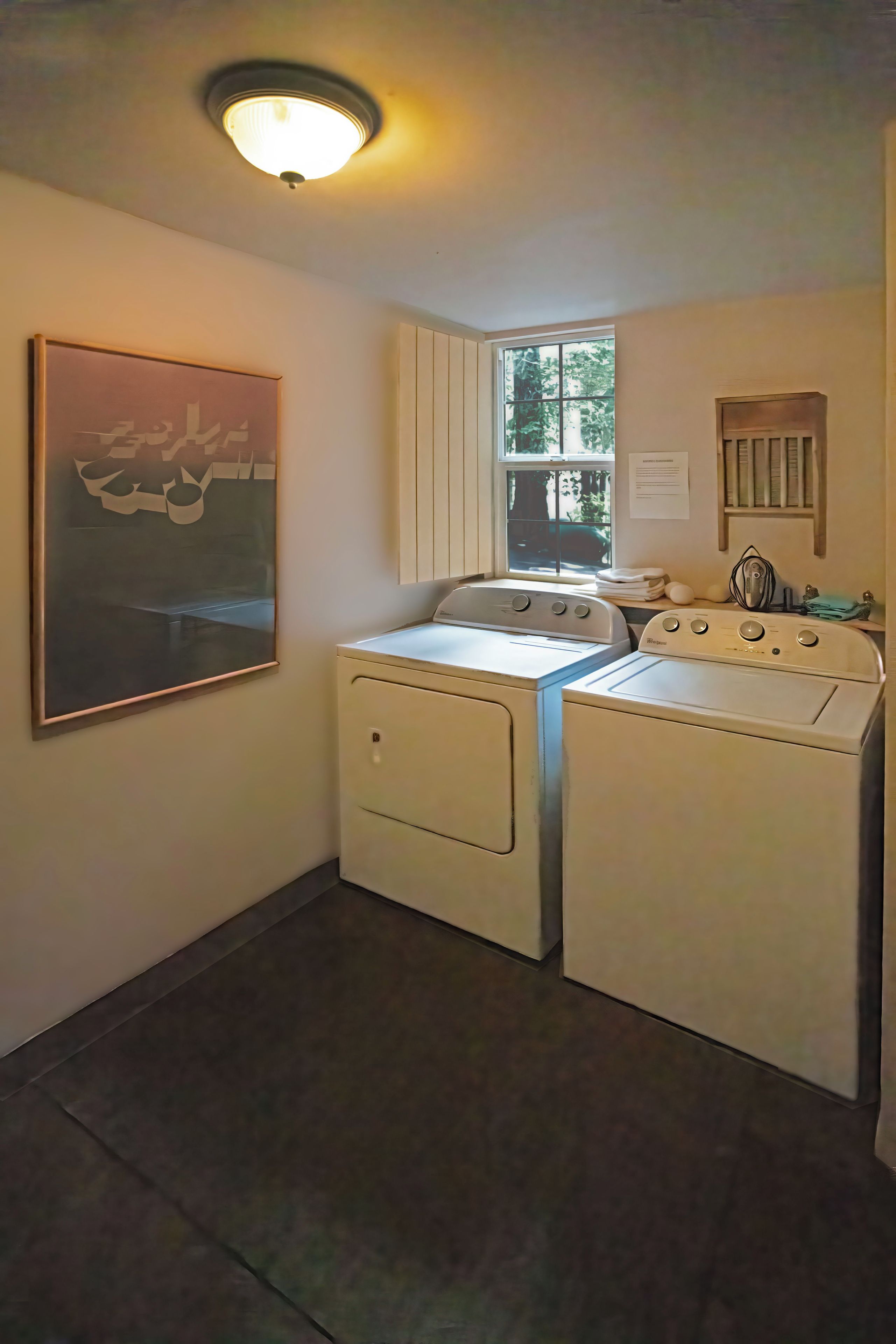 A laundry room with two washing machines, a ceiling light, a window, and a framed artwork on the wall.