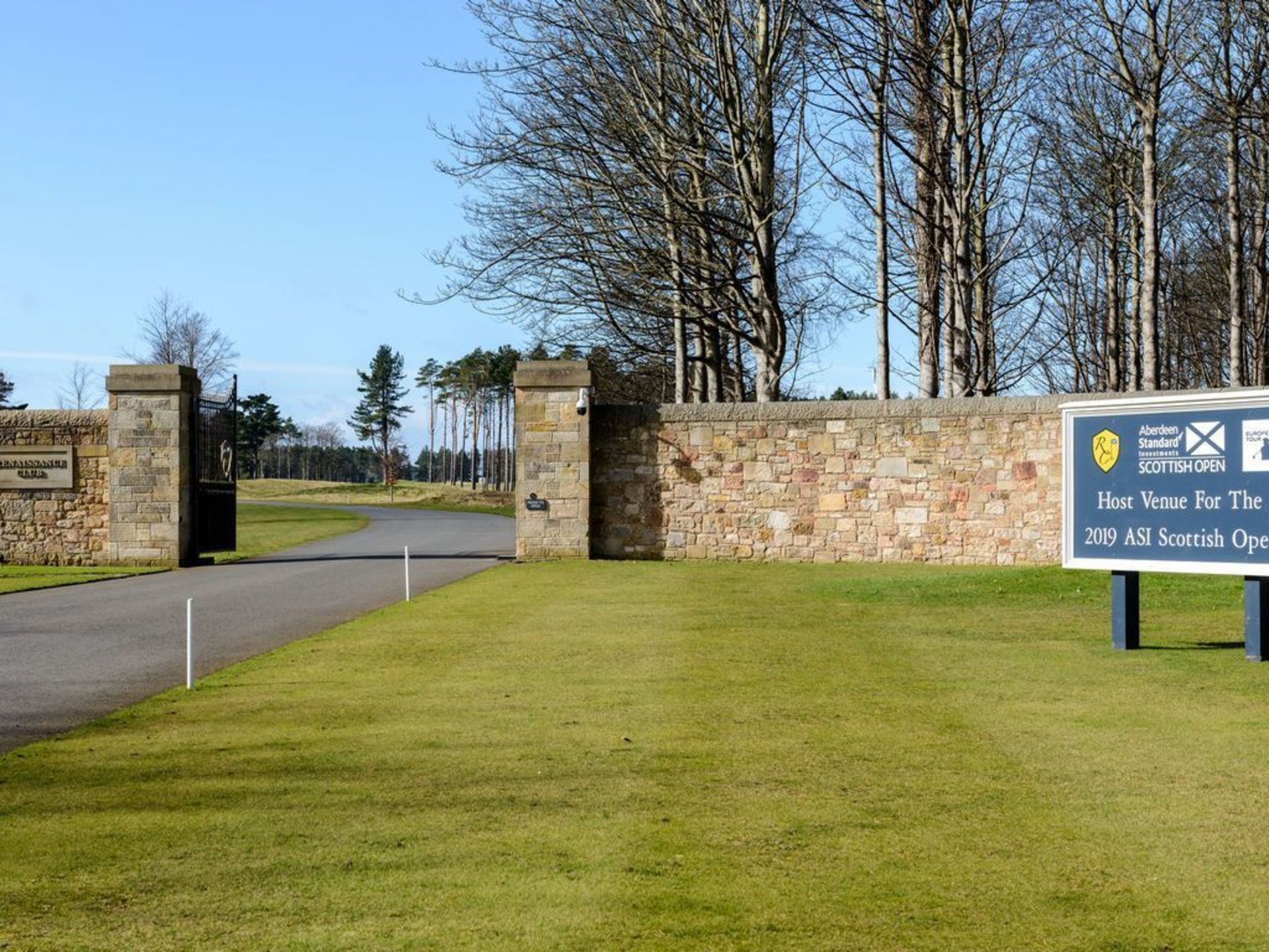 A stone wall entrance to a golf course with a sign indicating it is the host venue for the 2019 ASI Scottish Open.