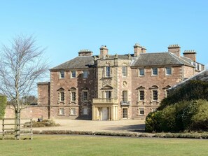 A large, historic stone building with multiple chimneys, a central entrance, and a well-maintained lawn.