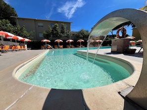 A swimming pool with a water feature, surrounded by lounge chairs and umbrellas, with a building in the background. - Residence le Rose (Bibbona)