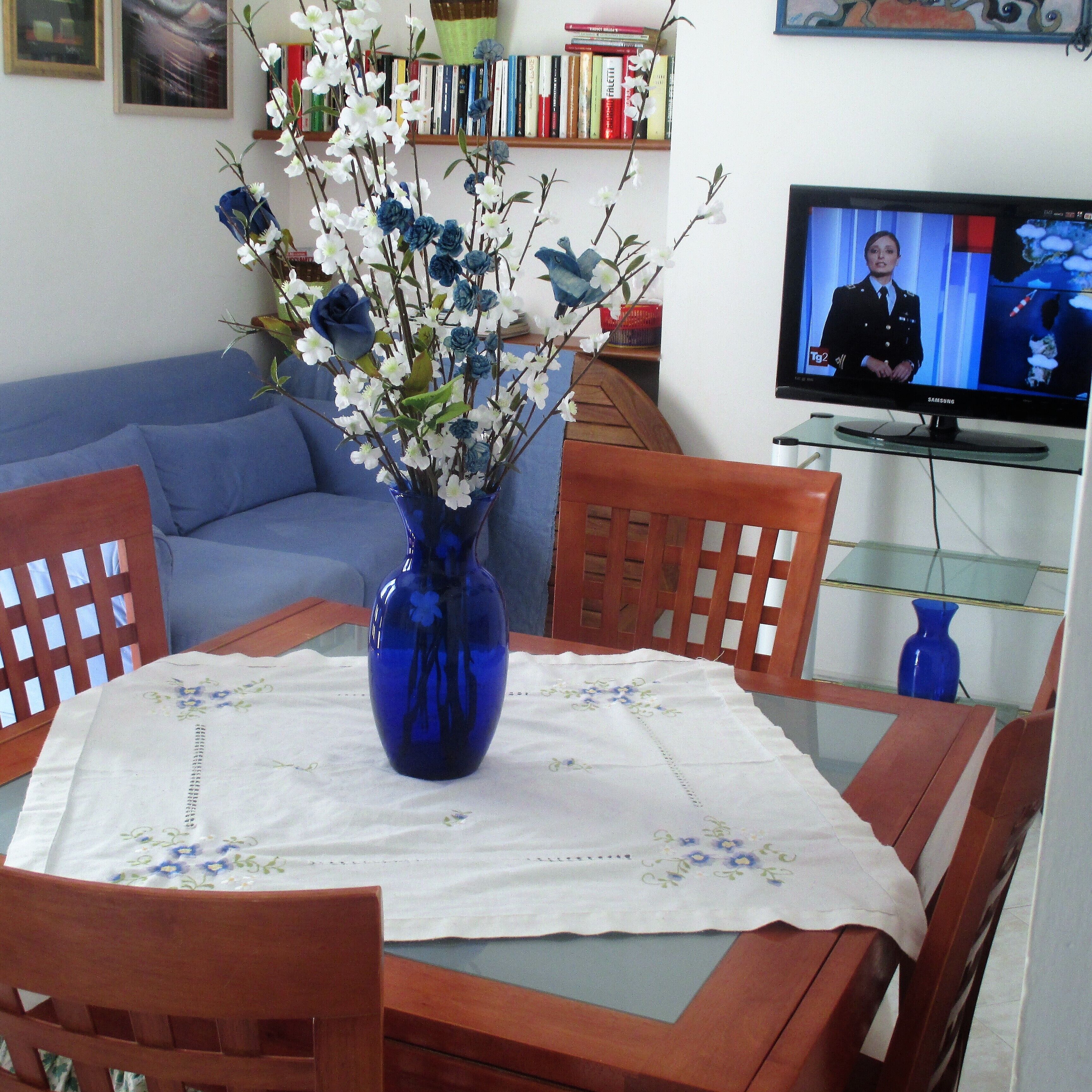 A living room with a blue vase on a table, a blue sofa, and a television on a stand.