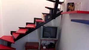 A compact room with a red and black staircase, a bed with a patterned bedspread, a television on a stand, and a shelf with books. - Portal Del Pinar (Bogotá)