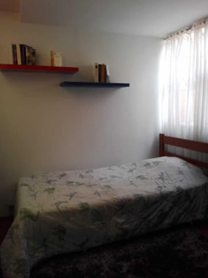 A small room with a single bed, a wooden headboard, two wall-mounted shelves with books, and a window with sheer curtains. - Portal Del Pinar (Bogotá)