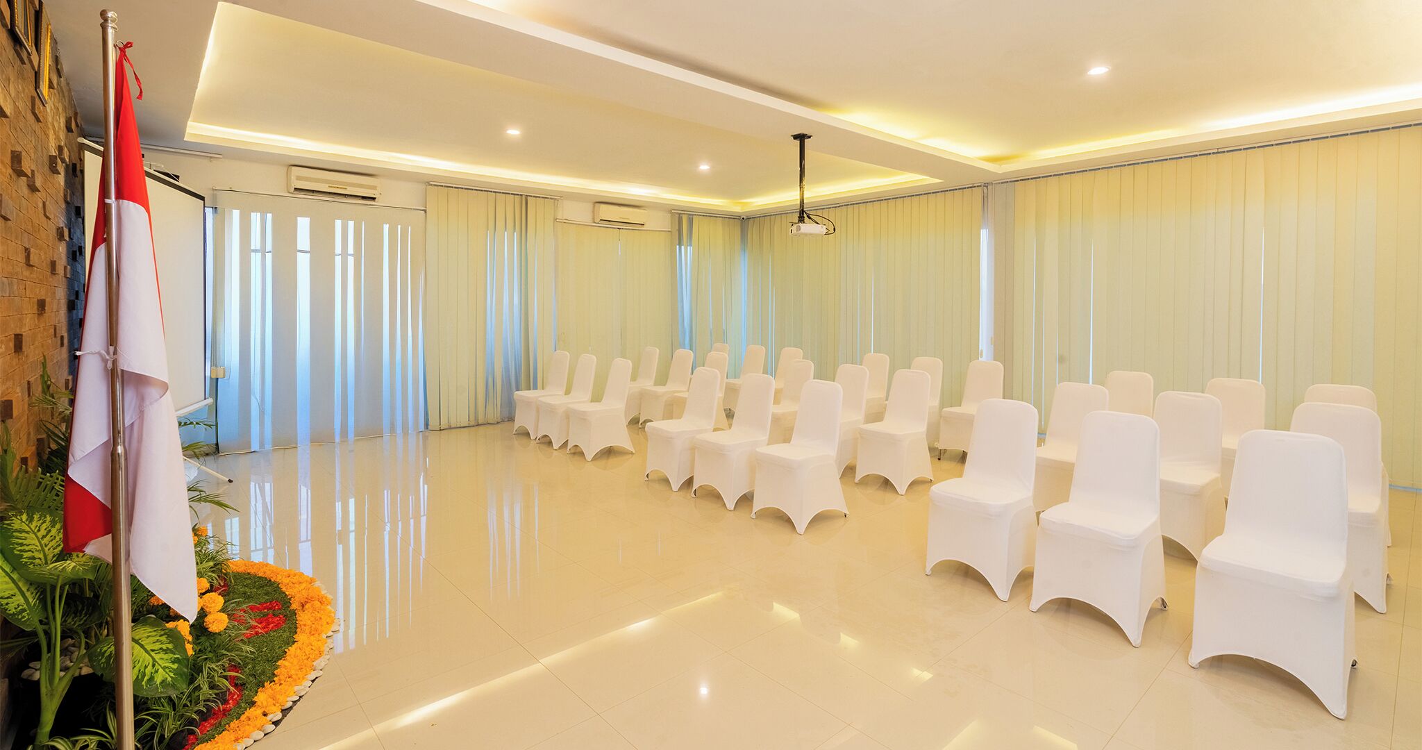 A spacious hall with rows of white chairs, air conditioning units, and a decorative plant arrangement.