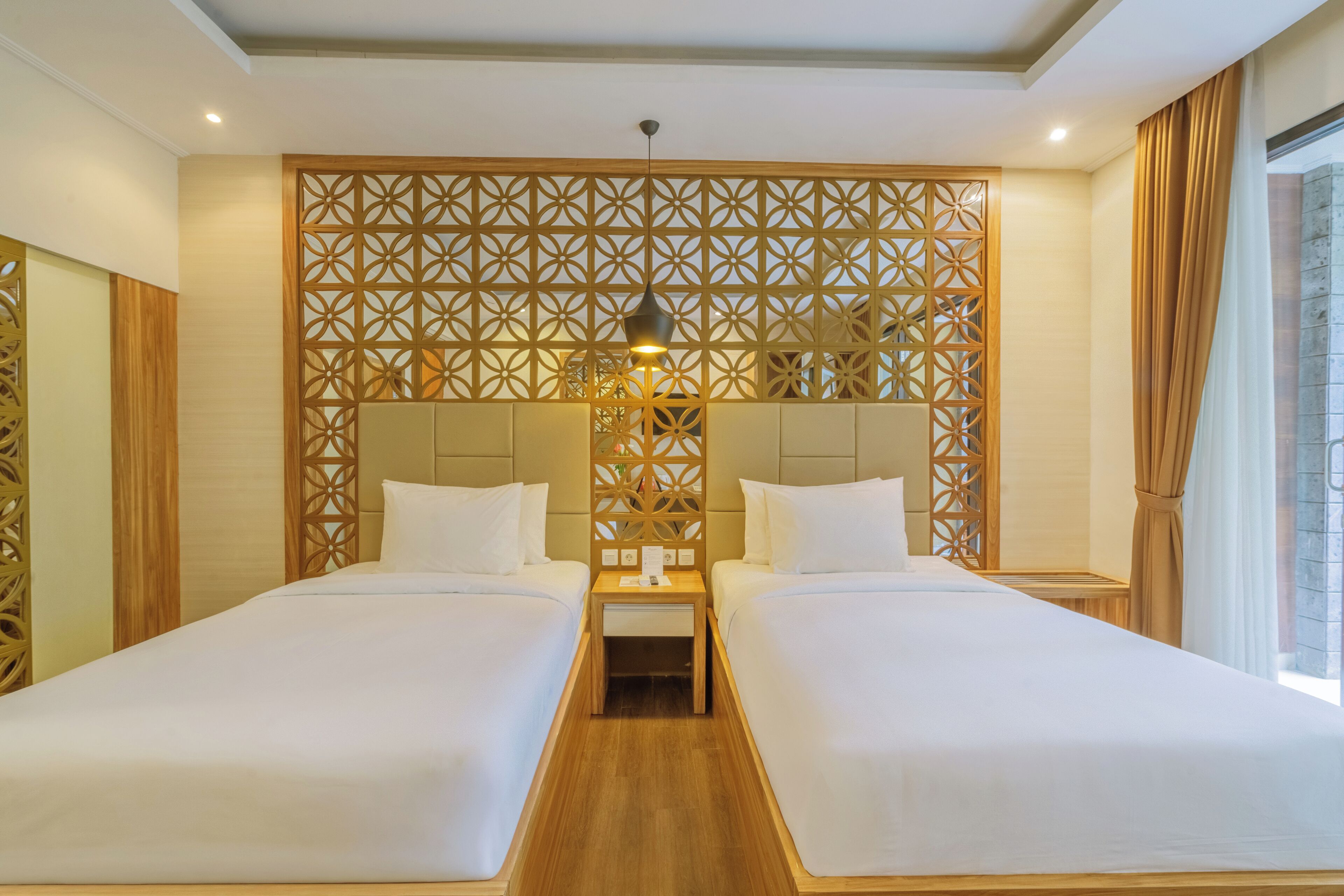 A hotel room with two beds, a decorative wooden partition, a small table, and a hanging light fixture.