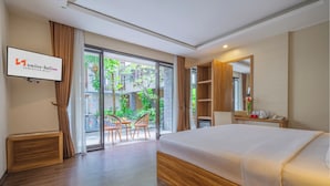 A modern hotel room with a large bed, a TV mounted on the wall, a wooden side table, and a sliding glass door leading to a balcony with outdoor seating. - Swiss-Belinn Seminyak (Denpasar)
