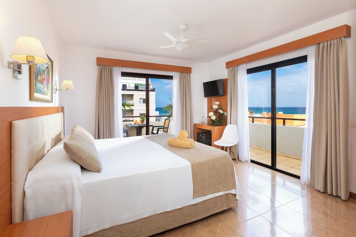A hotel room with a large bed, a desk with a chair, a TV, and a balcony with a view of the ocean.