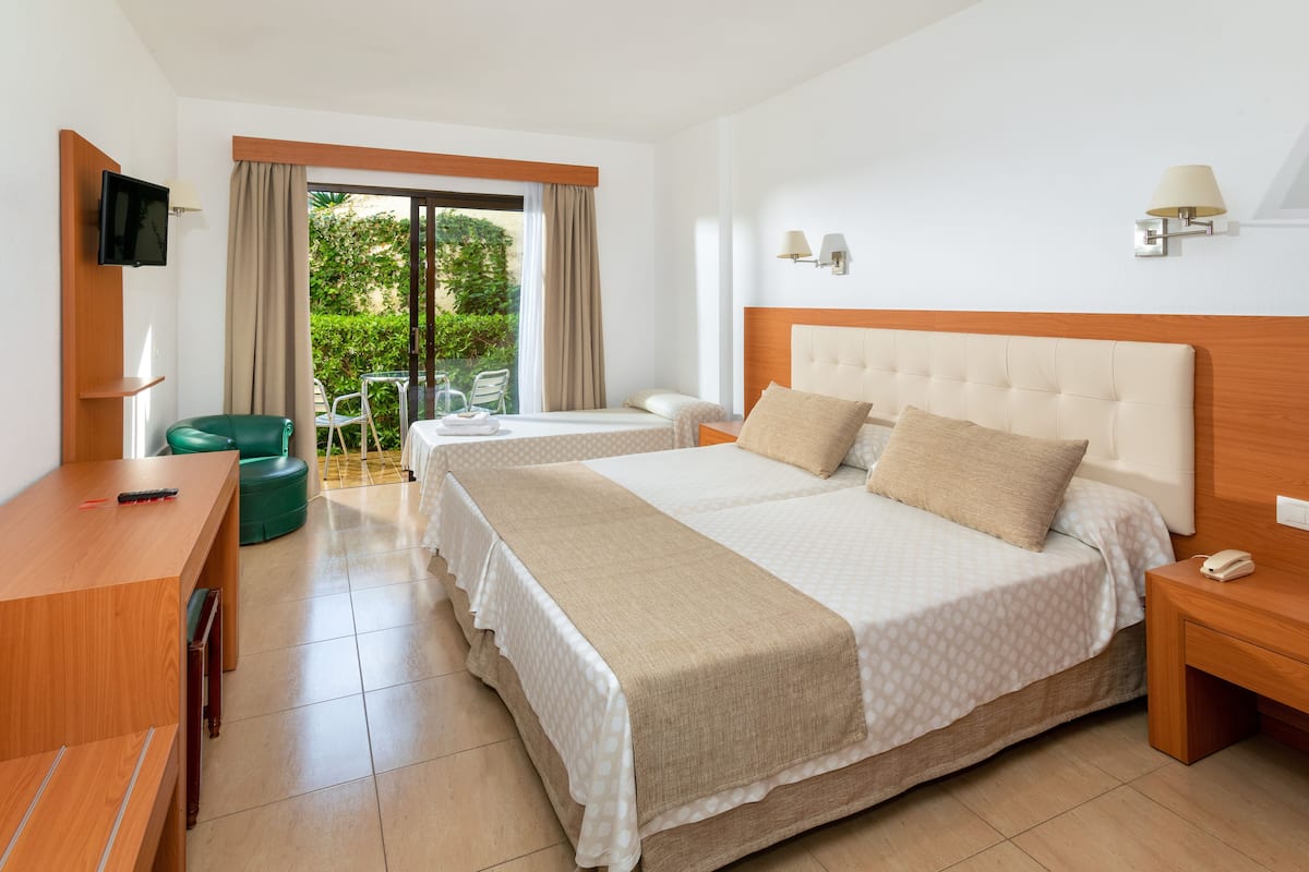 A hotel room with two beds, a desk, a chair, a TV, and a balcony with a view of greenery.