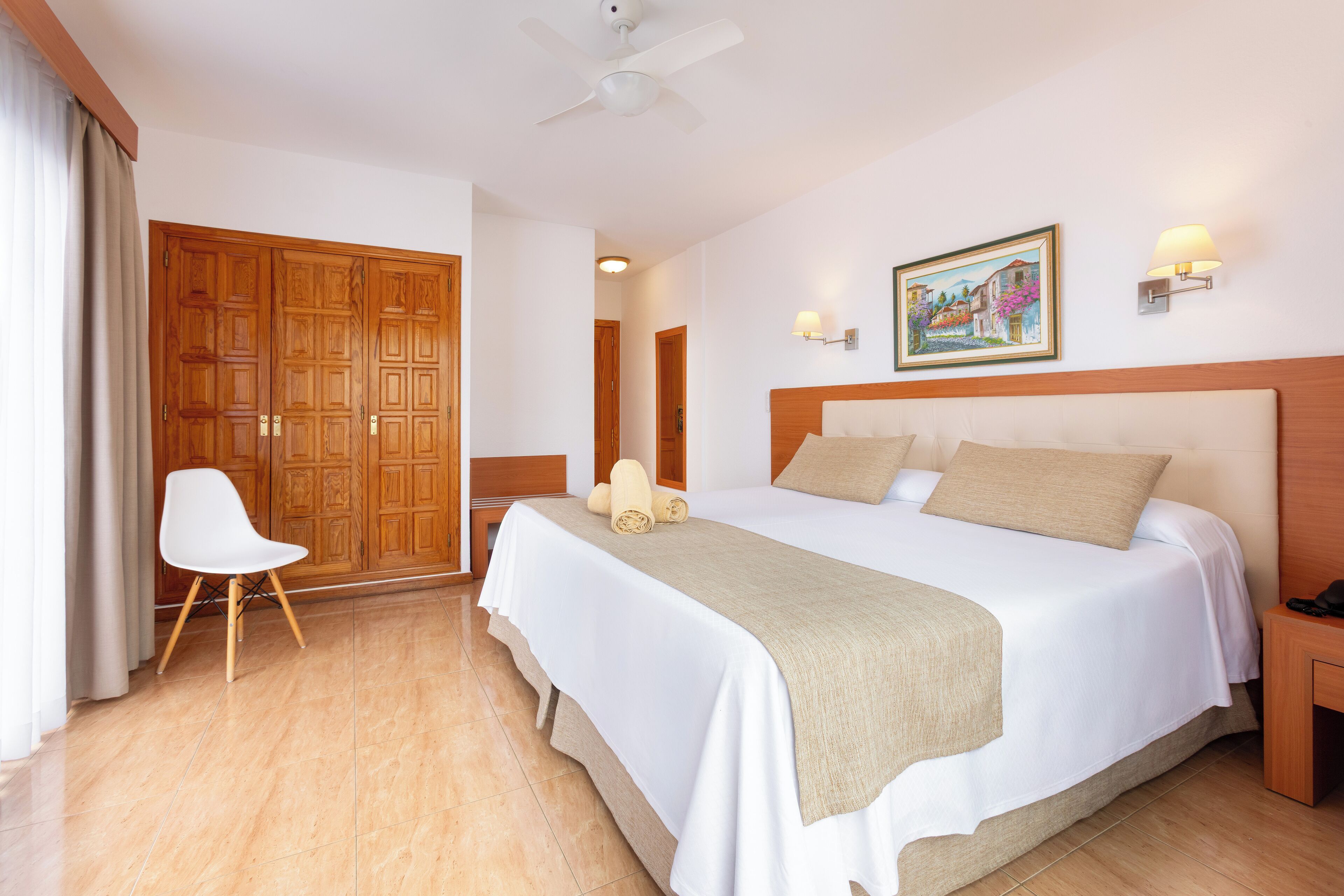 Una habitación de hotel con una cama grande, un armario de madera, una silla blanca y un cuadro en la pared.