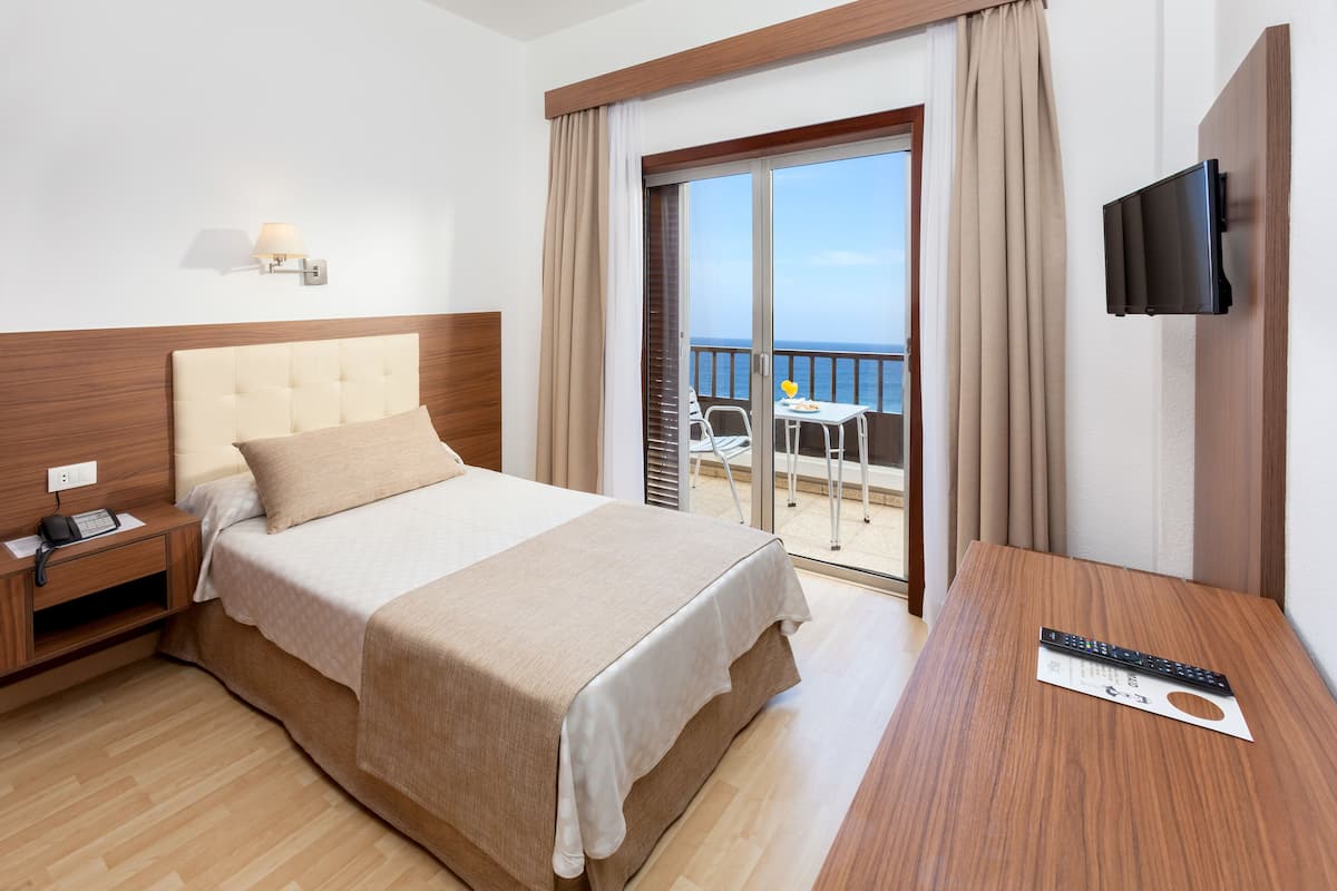 A hotel room with a bed, a bedside table with a phone, a TV mounted on the wall, and a balcony with a table and chairs.