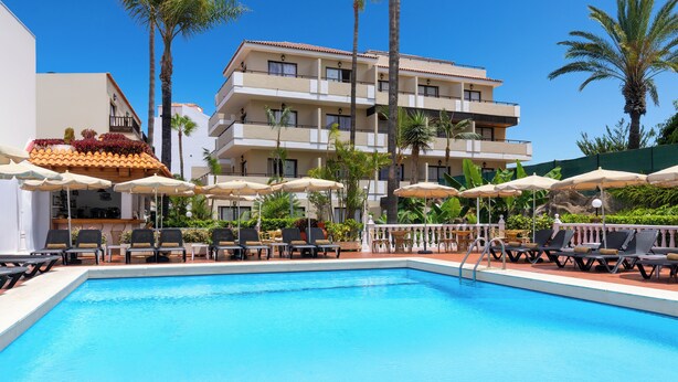 A swimming pool with lounge chairs and umbrellas, surrounded by palm trees and a multi-story building with balconies. - Hotel Don Manolito (Puerto de la Cruz)