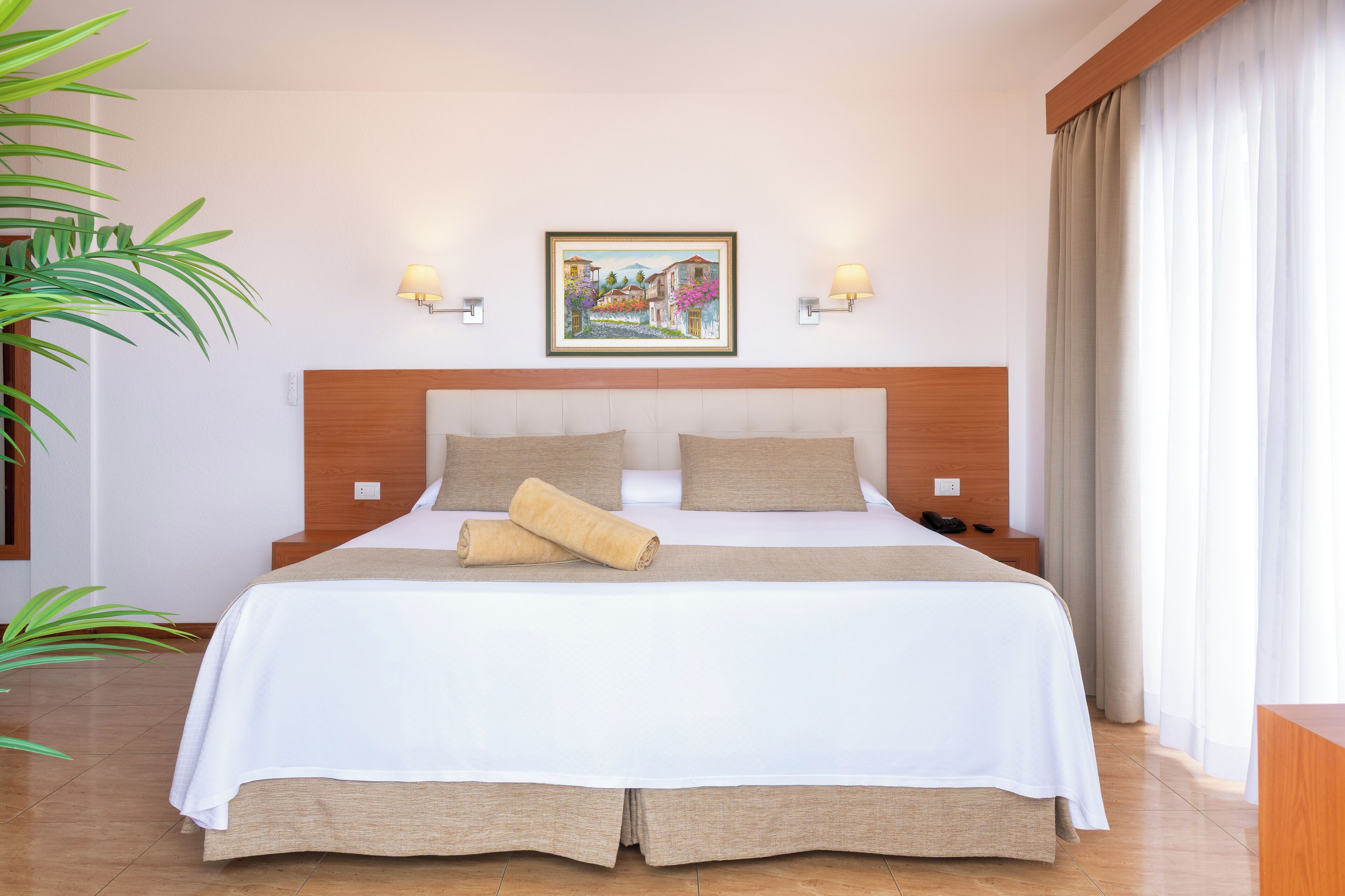 A neatly made bed with a white and beige bedspread, two pillows, and a rolled towel. There are two bedside tables with lamps, a painting on the wall, and a large window with curtains.