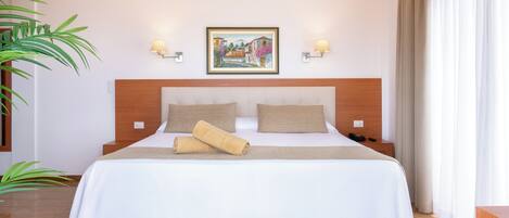 Una cama bien hecha con colcha blanca y color crema, dos almohadas y una toalla enrollada. Hay dos mesitas de noche con lámparas, un cuadro en la pared y un ventanal con cortinas.