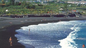 A beach with waves, a mountain in the background, and a town across the water. - Hotel Don Manolito (Puerto de la Cruz)