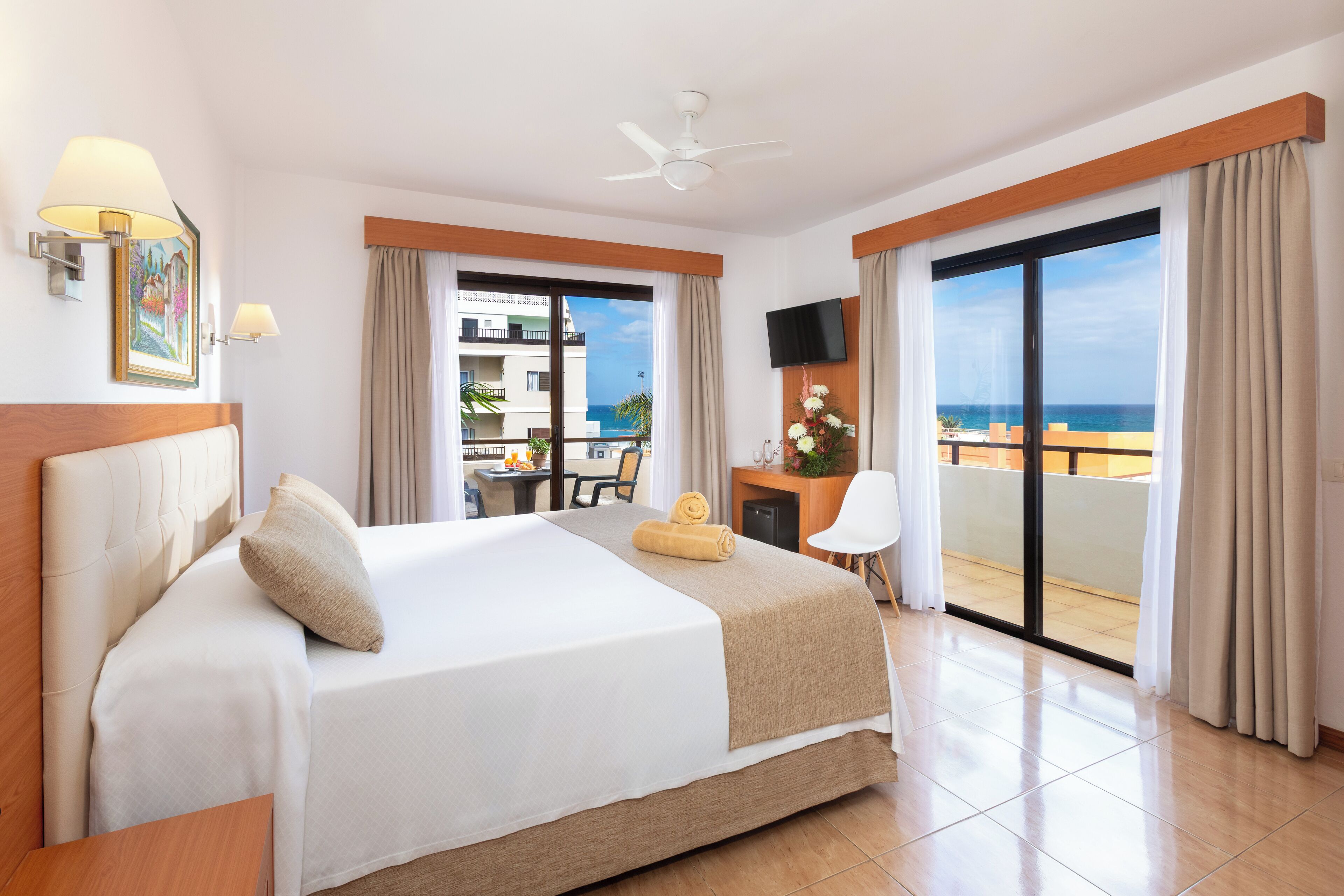 Una camera d'albergo con un letto grande, una scrivania con una sedia, una TV e un balcone vista mare.