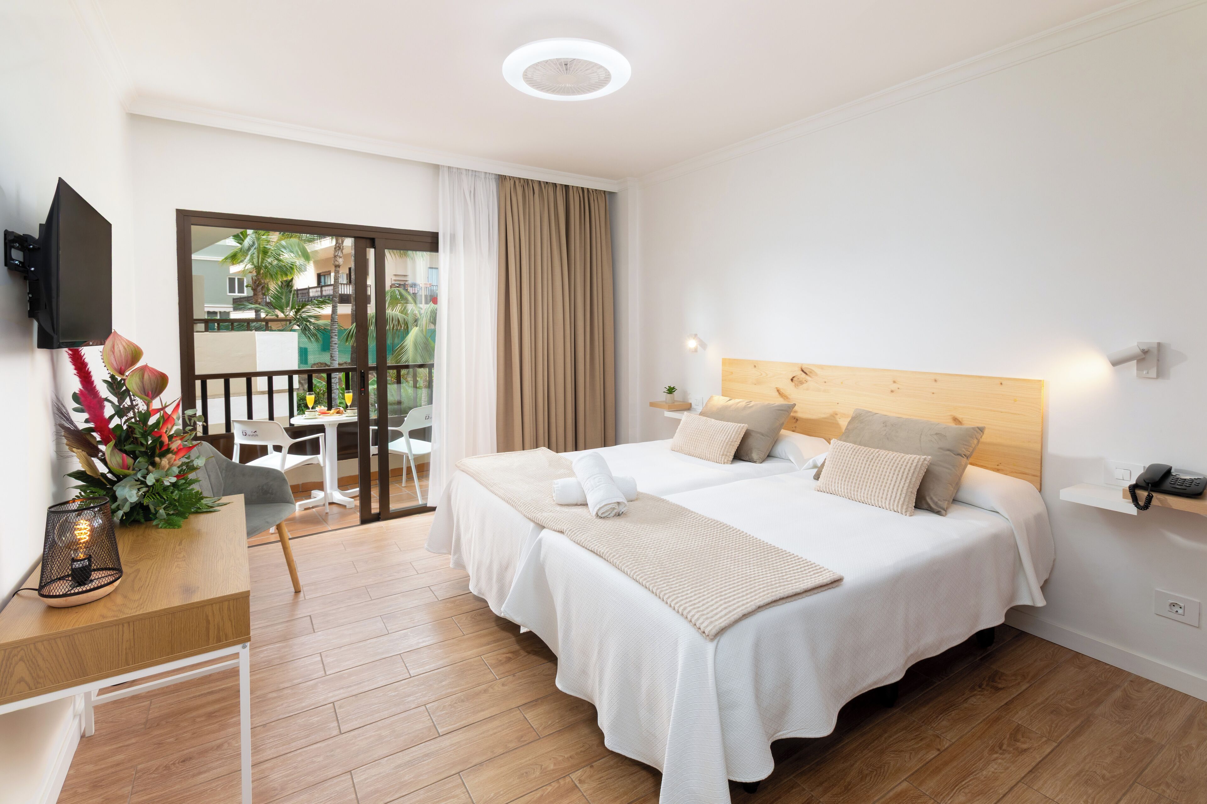 A hotel room with a bed, a desk with a lamp, a TV mounted on the wall, and a view of the outside balcony.