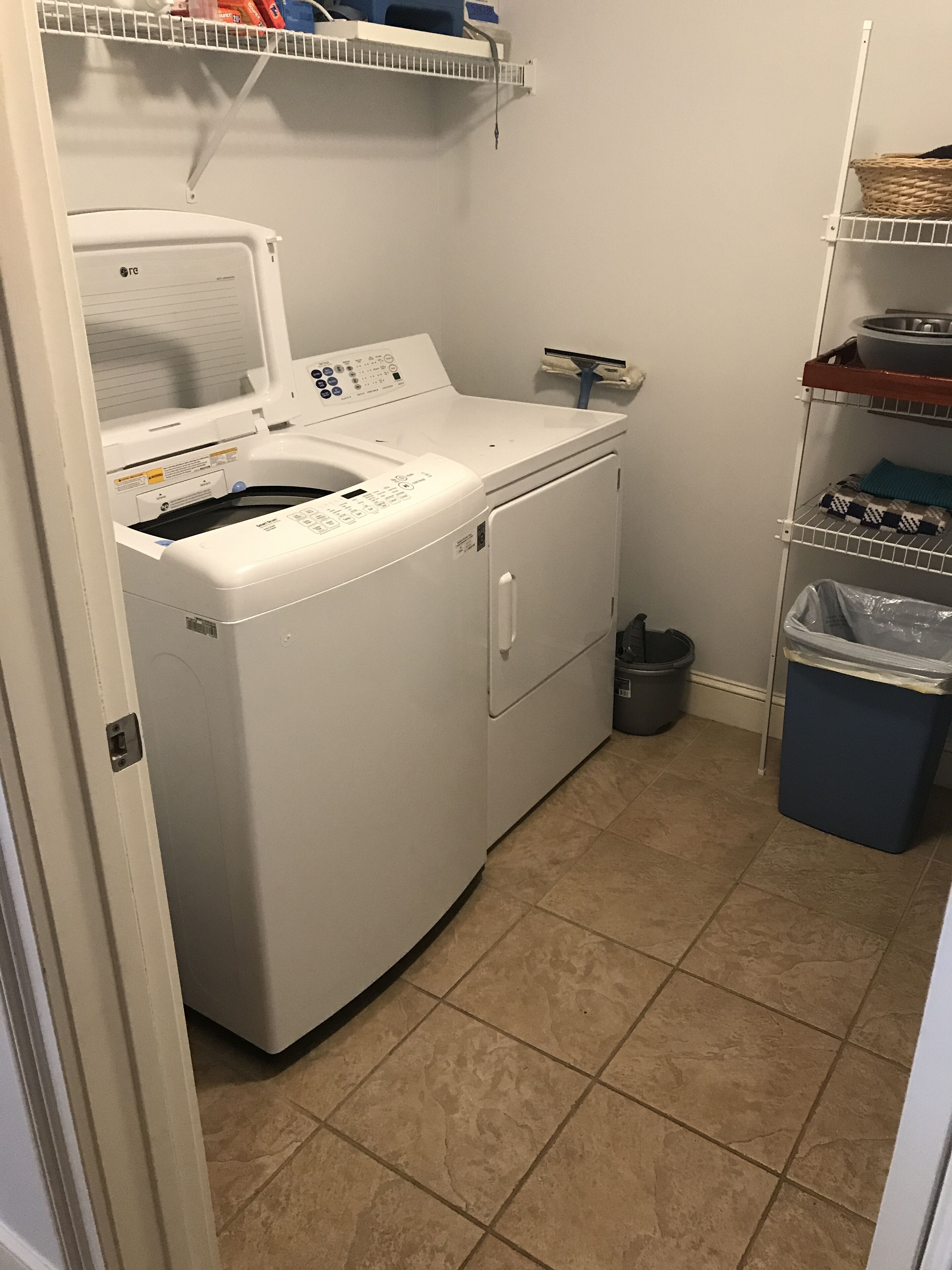 A laundry room with a front-loading washer and dryer, a trash can, and shelves with laundry supplies.