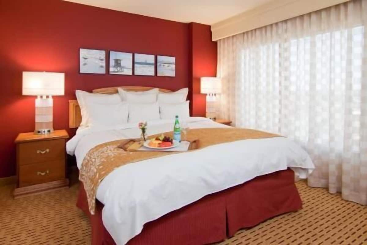 A hotel room with a large bed, two bedside tables, a nightstand, a wall-mounted lamp, a window with sheer curtains, and a small table with a plate of fruit.